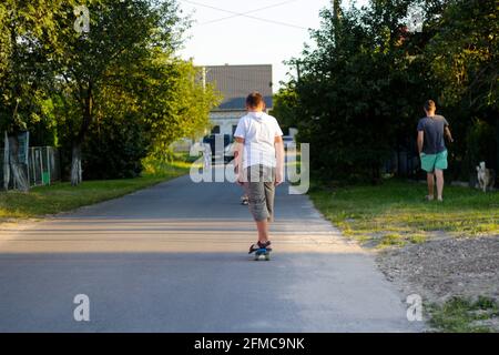 Redéfinit le jeune garçon heureux jouant sur le skateboard dans la rue. Homme marchant avec un chien en arrière-plan. Carace d'enfant de race blanche, planche de penny, pratique du skateboard Banque D'Images