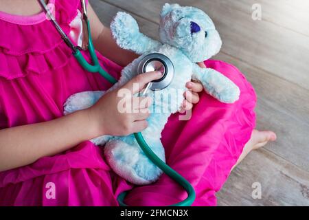 Enfant jouant médecin ou infirmière avec un ours en peluche à la maison. Une fille heureuse écoute un stéthoscope pour le jouet. Jeu de rôle de fille ludique. Banque D'Images