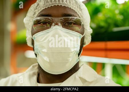 Portrait d'un jeune spécialiste de la culture afro-américaine sérieux en masque, lunettes et casquette debout contre le plateau vertical de la ferme Banque D'Images