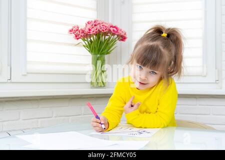 une petite fille s'assoit à une table dans une pièce blanche et dessine. Banque D'Images