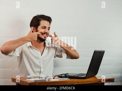 Jeune homme caucasien faisant un geste de sourire forcé tout en travaillant dans bureau Banque D'Images