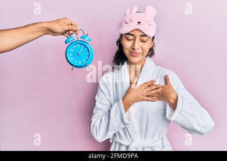 Adolescente hispanique avec bretelles dentaires portant un masque de sommeil et une robe avec réveil près d'elle souriante avec les mains sur la poitrine, les yeux fermés avec le sourire Banque D'Images