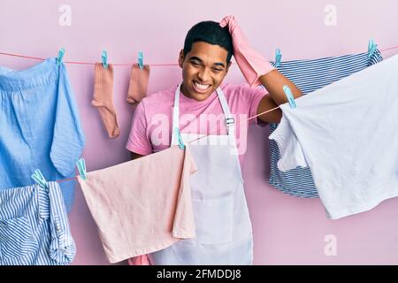 Jeune homme hispanique beau portant un tablier plus propre tenant des vêtements sur la corde à linge confondre et se poser des questions. Incertain avec le doute, penser avec humour Banque D'Images
