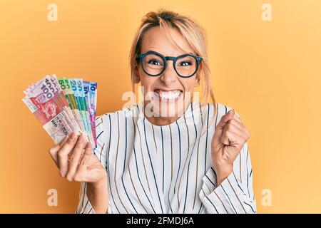 Belle femme blonde tenant des billets de hong kong dollars criant fièrement, célébrant la victoire et le succès très excité avec le bras relevé Banque D'Images