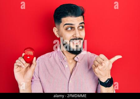 Beau homme avec la barbe tenant l'anneau d'engagement pour le pointage de proposition le pouce jusqu'au côté souriant heureux avec la bouche ouverte Banque D'Images