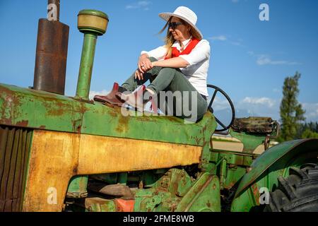 Femme urbaine vêtue de vêtements décontractés et assise sur un vieux tracteur contre le ciel bleu dans les terres agricoles Banque D'Images