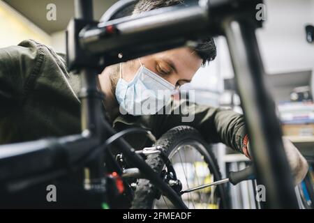 Atelier de vélo le réparateur travaille dans l'atelier d'entretien et de réparation de vélo pendant la quarantaine du coronavirus portant un écran facial et des gants, nouvelle norme. Mécanicien Banque D'Images