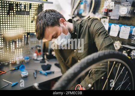 Atelier de vélo le réparateur travaille dans l'atelier d'entretien et de réparation de vélo pendant la quarantaine du coronavirus portant un écran facial et des gants, nouvelle norme. Mécanicien Banque D'Images