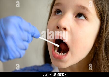 Test PCR COVID-19 et petit visage en gros plan, petite fille ouvre la bouche pour le test du coronavirus. Le médecin ou l'infirmière tient un écouvillon pour l'échantillon de salive d'un enfant mignon Banque D'Images