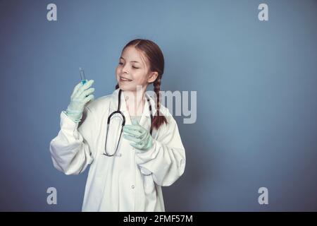 belle, fraîche jeune fille vêtue comme femme médecin avec stéthoscope et seringue et gants en caoutchouc posant devant un fond bleu Banque D'Images