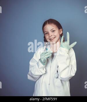 belle, fraîche jeune fille vêtue comme femme médecin avec stéthoscope et seringue et gants en caoutchouc posant devant un fond bleu Banque D'Images