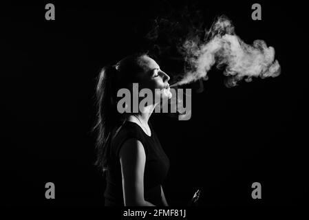 La brunette fume une cigarette électronique sur fond noir, dégage une fumée épaisse de sa bouche. Monochrome. Portrait d'une femme fumant un singe Banque D'Images