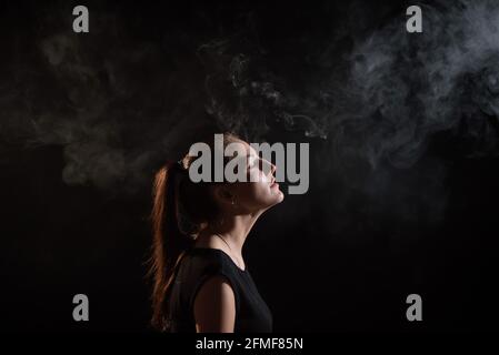 La brunette fume une cigarette électronique sur fond noir, dégage une fumée épaisse de sa bouche. Portrait d'une femme fumant un singe, planant. Banque D'Images