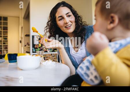 Femme parlant avec son bébé tout en le nourrissant à la maison. Une mère qui s'occupe de nourrir un tout-petit en congé de maternité. Banque D'Images