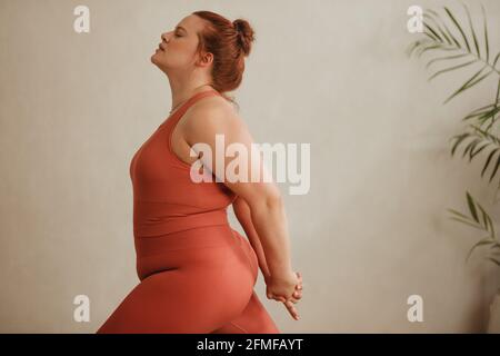 Vue latérale d'une femme forte qui fait de l'exercice. Plus grande taille femme en vêtements de sport faisant quelques exercices d'étirement avec la main clased derrière. Banque D'Images
