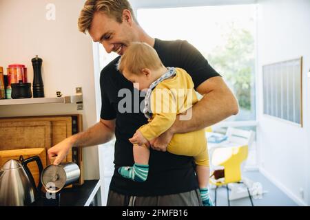 Père transportant son bébé et préparant le café dans la cuisine. Homme en congé de paternité avec son tout-petit debout dans la cuisine et faire du café. Banque D'Images