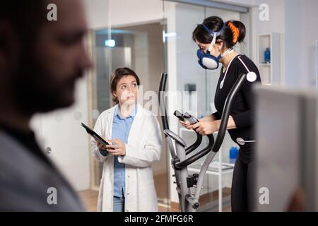 Sportswoman courant avec masque sur entraîneur croisé avec des électrodes fixées au corps. Médecin utilisant un ordinateur tablette et contrôle les données EKG affichées sur les moniteurs de laboratoire, discutant avec le patient. Banque D'Images