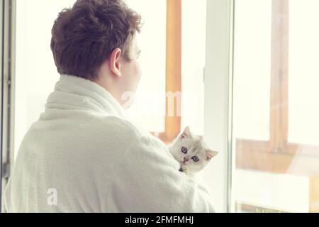 Un jeune homme du caucase boit du café près de la fenêtre. Il tient un chat. Tôt le matin, soleil. Banque D'Images