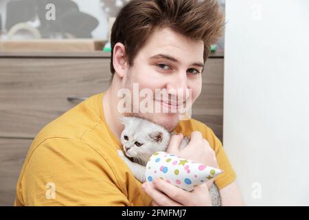 Triste chaton blanc dans un chapeau de fête dans les bras d'un jeune homme. Concept anniversaire et vacances. Banque D'Images