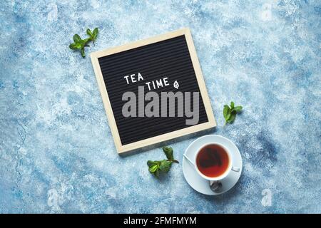 Temps de thé. Tableau de lettres noir avec texte sur table bleue avec tasse de thé en verre et feuilles de menthe. Vue de dessus Banque D'Images