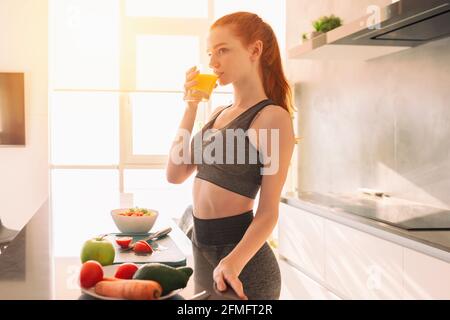 Une jeune femme athlétique aux cheveux rouges dans la cuisine boit un verre de jus de fruits centrifugé Banque D'Images