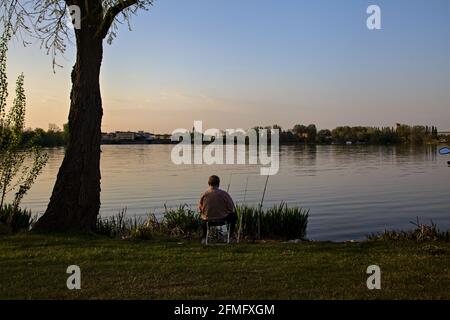 Vieux pêcheur assis sur la rive d'un lac à coucher de soleil Banque D'Images