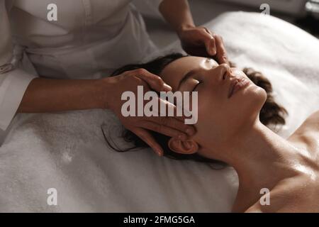 Jeune femme bénéficiant d'un massage au spa. Esthéticienne frottant les temples de client, fille souriante se relaxant pendant la thérapie de massage myofascial à la beauté Banque D'Images