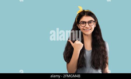 Vérifiez ceci. Joyeuse fille indienne ado pointant de côté avec le pouce vers le haut à l'espace vide sur fond bleu, conception de bannière Banque D'Images