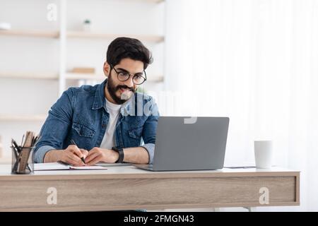 Homme d'affaires arabe prenant des notes tout en travaillant sur un ordinateur portable au bureau à domicile, espace vide Banque D'Images