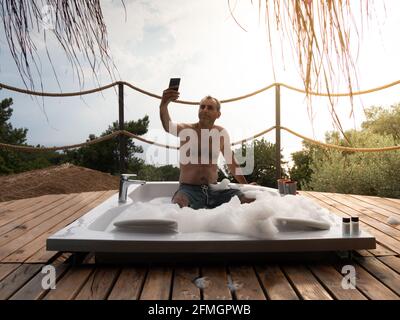 Un bel homme adulte prenant le selfie dans un bain à remous dans un luxueux spa exotique hôtel balcon avec vue sur la mer et le coucher du soleil. Banque D'Images