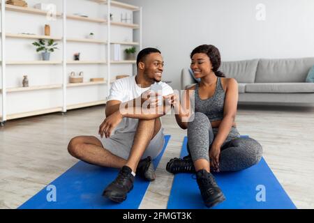 Jeune couple noir assis sur des tapis de yoga, prenant une pause de l'entraînement à la maison, regardant les conseils sportifs en ligne sur smartphone Banque D'Images