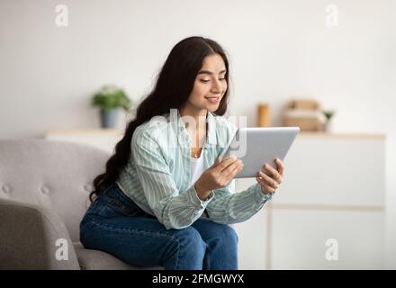 Gentille dame indienne assise dans un fauteuil avec une tablette, étudiant à distance ou ayant une réunion en ligne à la maison Banque D'Images