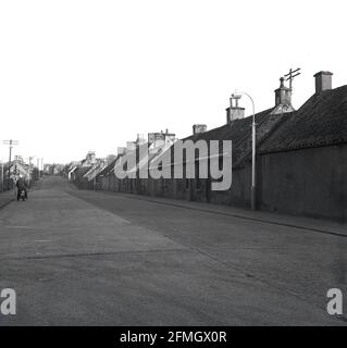 Années 1960, historique, un motocycliste sur la rue principale vide à travers le village de Cairneyhill, montrant de vieux, simples maisons de tisserand, certains abandonnés, Fife, Écosse. Banque D'Images