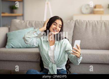 Belle femme indienne portant un casque, choisissant une liste de lecture musicale sur un appareil mobile, écoutant votre chanson préférée à la maison Banque D'Images