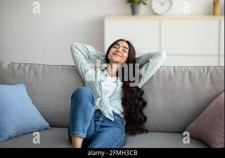 Week-end de détente à la maison. Jeune femme indienne décontractée assise sur un canapé, tenant les mains derrière sa tête, fermant les yeux Banque D'Images