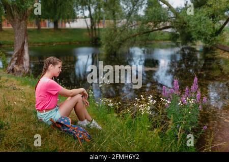 Une fille caucasienne aux taches de rousseur et dans un t-shirt rose se trouve sur l'herbe. Une jeune étudiante sourit et apprécie le beau temps d'été. Banque D'Images