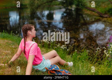 Une fille caucasienne aux taches de rousseur et dans un t-shirt rose se trouve sur l'herbe. Une jeune étudiante sourit et apprécie le beau temps d'été. Banque D'Images