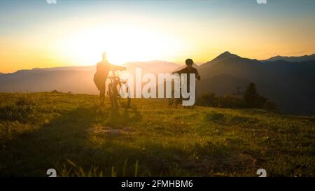 Deux silhouettes poussant un VTT, à travers la prairie au sommet de la colline pendant le coucher du soleil Banque D'Images