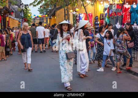 De jeunes femmes vietnamiennes vêtues de chapeaux coniques traditionnels et de robes traditionnelles ao dai marchent à travers la vieille ville, Hoi an, Vietnam Banque D'Images