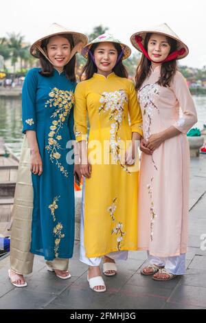 Trois jolies femmes vietnamiennes portant des chapeaux coniques et des robes AO dai traditionnelles posent pour la caméra par la vieille ville au bord de la rivière, Hoi an, Vietnam Banque D'Images