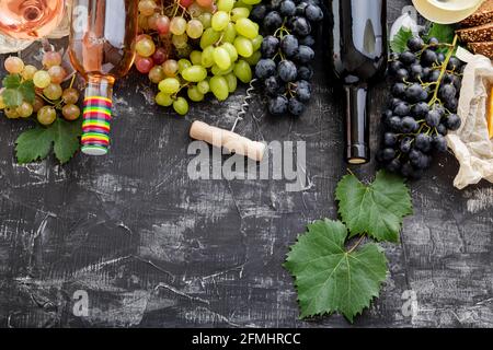 Vin rouge rose en bouteilles de fromage, raisin blanc rose et noir avec plante de vigne sur fond de béton foncé. Assortiment différents types de vin et de raisin Banque D'Images