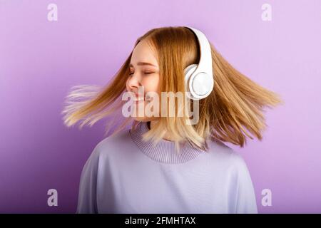 Belle femme souriante dansant dans des écouteurs avec la coiffure de cheveux blond volant. Une adolescente aime écouter de la musique en se déplaçant dans un casque sans fil Banque D'Images