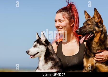 Content athlète féminine avec piercing embrassant Husky sibérien et allemand Shepherd tout en regardant à l'abri de la lumière du soleil Banque D'Images