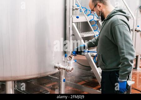 Vue latérale de l'ouvrier de la récolte dans la vanne de lavage du masque de réservoir de métal au travail dans la société de brassage pendant la pandémie Banque D'Images