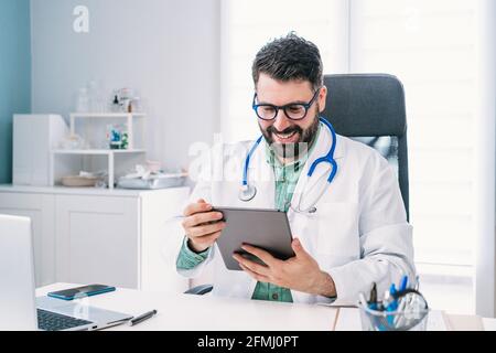 Joyeux médecin de sexe masculin d'âge moyen en lunettes et uniforme avec tablette pendant le chat vidéo à l'hôpital Banque D'Images