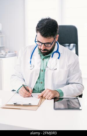 Barbu mature homme médecin en uniforme avec stéthoscope prenant des notes sur une feuille de papier pendant le travail en clinique Banque D'Images