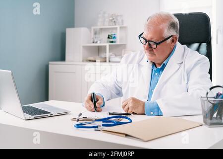 Homme âgé médecin en uniforme avec stéthoscope prenant des notes sur feuille de papier pendant le travail en clinique Banque D'Images