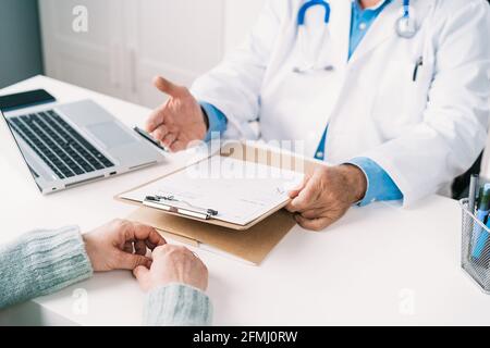 Un médecin masculin âgé non reconnaissable a été mis en culture sous forme uniforme sur la planchette à pince de la table contre une femme méconnaissable pendant le contrôle clinique Banque D'Images