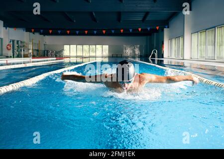 Un grand nageur masculin dans une casquette de bain effectuant un mouvement de papillon pendant faites de l'exercice dans la piscine avec de l'eau bleue Banque D'Images