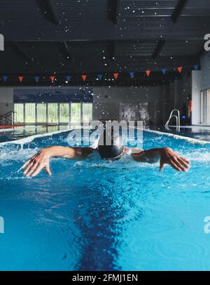 Un grand nageur masculin dans une casquette de bain effectuant un mouvement de papillon pendant faites de l'exercice dans la piscine avec de l'eau bleue Banque D'Images